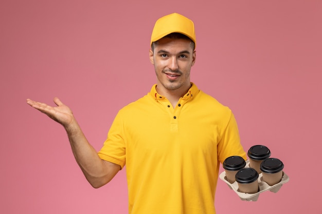 Vue avant de courrier masculin en uniforme jaune souriant et tenant des tasses de café de livraison sur le bureau rose