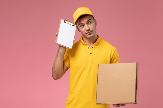 Vue avant de courrier masculin en uniforme jaune tenant le bloc-notes avec boîte de livraison de nourriture pensant sur le fond rose