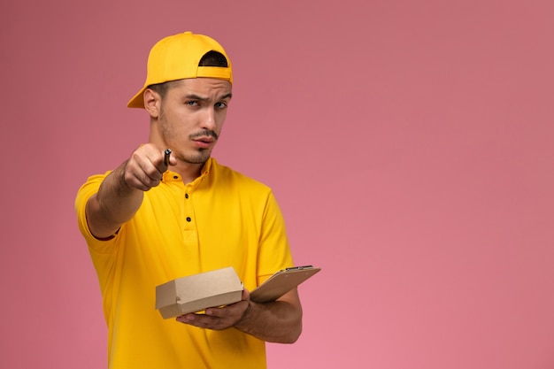 Vue avant de courrier masculin en uniforme jaune tenant le bloc-notes et petit paquet de nourriture écrit des notes sur un bureau rose.