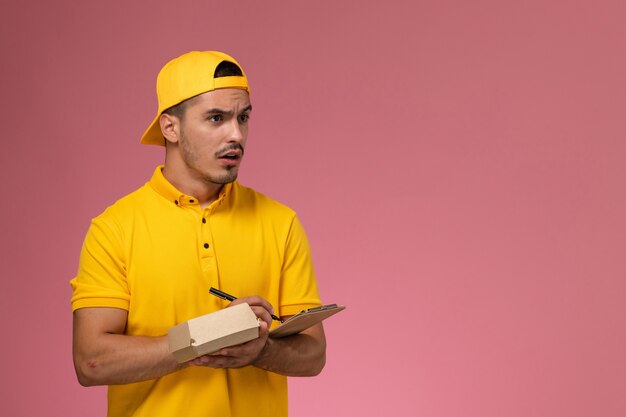 Vue avant de courrier masculin en uniforme jaune tenant le bloc-notes et petit paquet de nourriture écrit des notes sur le fond rose.