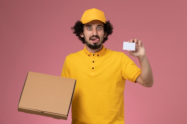 Vue avant de courrier masculin en uniforme jaune tenant la boîte et la carte de livraison de nourriture sur le mur rose