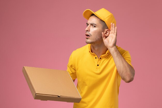 Vue avant de courrier masculin en uniforme jaune tenant la boîte de livraison de nourriture et en essayant d'entendre sur le fond rose