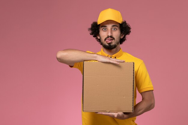 Vue avant de courrier masculin en uniforme jaune tenant la boîte de livraison de nourriture sur le mur rose clair