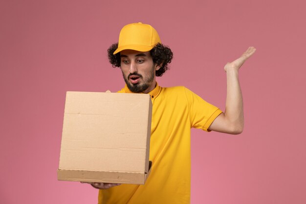 Vue avant de courrier masculin en uniforme jaune tenant et ouvrant la boîte de livraison de nourriture sur le mur rose clair