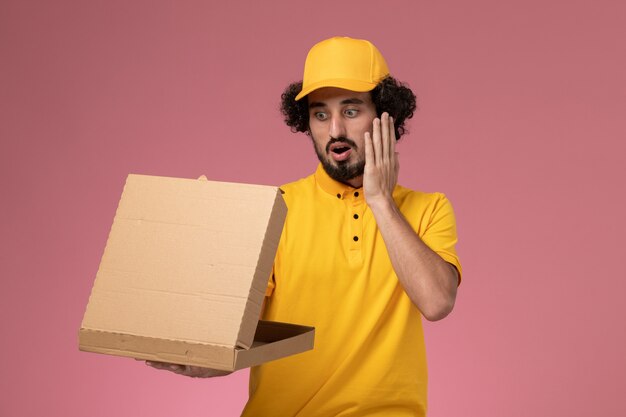 Vue avant de courrier masculin en uniforme jaune tenant et ouvrant la boîte de livraison de nourriture sur le mur rose clair