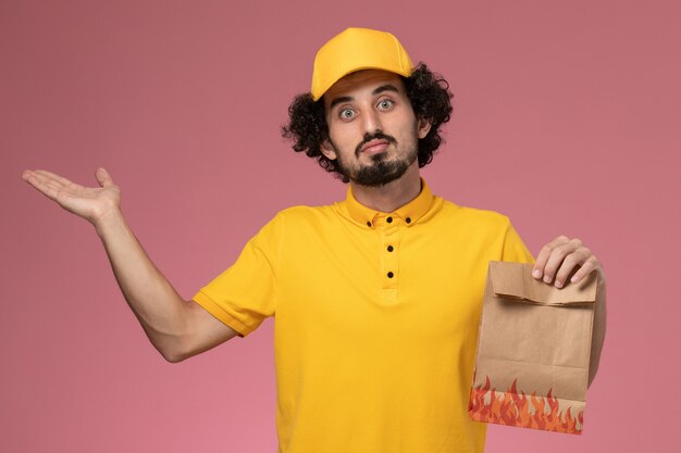 Vue avant de courrier masculin en uniforme jaune tenant un paquet de papier alimentaire sur un mur rose clair