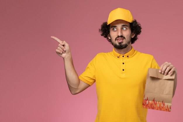 Vue avant de courrier masculin en uniforme jaune tenant un paquet de papier alimentaire sur un mur rose clair