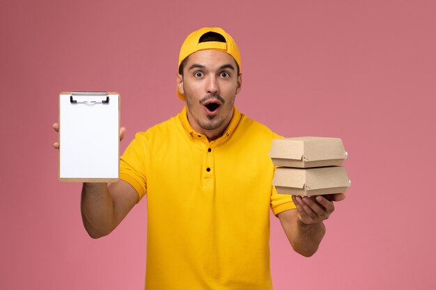 Vue avant de courrier masculin en uniforme jaune tenant de petits paquets de nourriture et petit bloc-notes sur un bureau rose