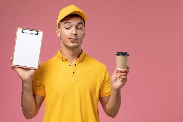 Vue avant de courrier masculin en uniforme jaune tenant la tasse de café de livraison et le bloc-notes sur le fond rose