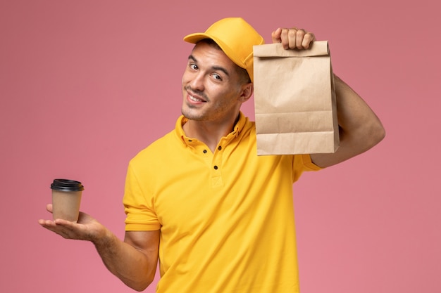 Vue avant de courrier masculin en uniforme jaune tenant la tasse de café de livraison et le paquet de nourriture sur le bureau rose clair