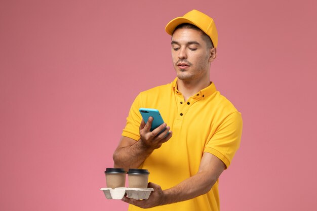 Vue avant de courrier masculin en uniforme jaune tenant des tasses de café de livraison à l'aide de son téléphone sur le bureau rose