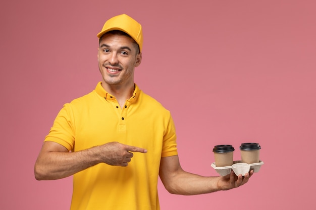 Vue avant de courrier masculin en uniforme jaune tenant des tasses de café de livraison sur un bureau rose clair