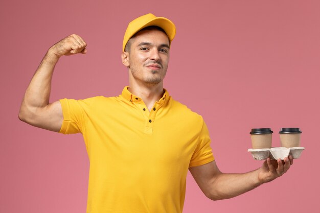 Vue avant de courrier masculin en uniforme jaune tenant des tasses de café de livraison et fléchissant sur fond rose clair