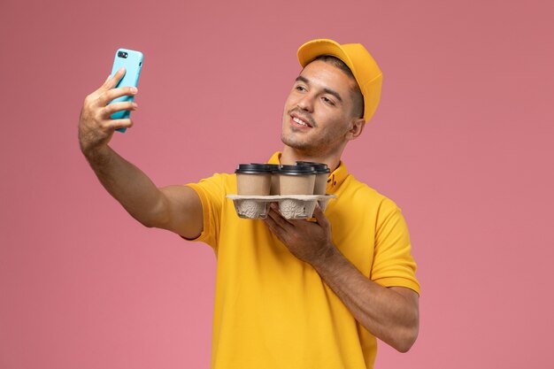 Vue avant de courrier masculin en uniforme jaune tenant des tasses de café de livraison prenant selfie sur bureau rose