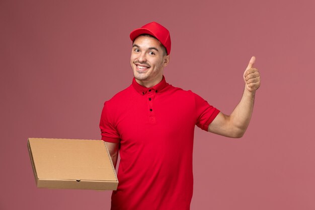 Vue avant de courrier masculin en uniforme rouge et cape tenant la boîte de livraison de nourriture sur l'emploi de travailleur de mur rose