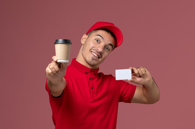 Vue avant de courrier masculin en uniforme rouge tenant la tasse de café de livraison et carte blanche sur le mur rose