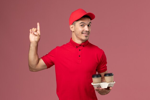 Vue avant de courrier masculin en uniforme rouge tenant des tasses de café de livraison marron avec un léger sourire sur le mur rose