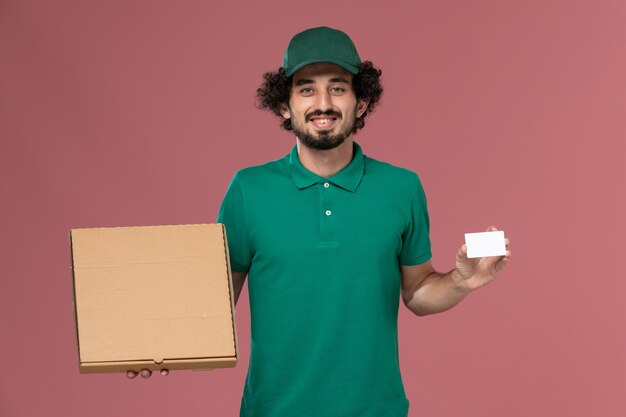 Vue avant de courrier masculin en uniforme vert et cape tenant la boîte de nourriture de livraison et la carte sur le travail de travail de livraison uniforme de service de fond rose