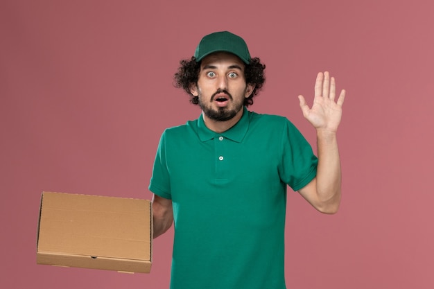 Vue avant de courrier masculin en uniforme vert et cape tenant la boîte de nourriture posant avec expression surprise sur la livraison uniforme de travail de bureau rose