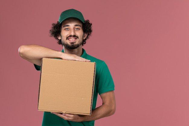 Vue avant de courrier masculin en uniforme vert et cape tenant la boîte de nourriture avec sourire sur la livraison uniforme de service fond rose
