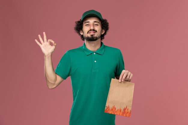 Vue avant de courrier masculin en uniforme vert et cape tenant un paquet de papier alimentaire sur le travail de livraison uniforme de travailleur de service de fond rose