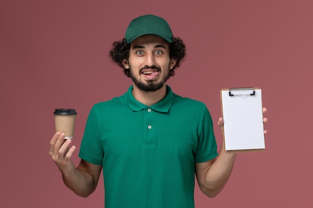 Vue avant de courrier masculin en uniforme vert et cape tenant la tasse de café de livraison avec bloc-notes sur fond rose clair service de livraison uniforme