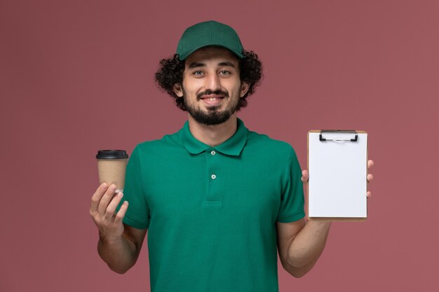 Vue avant de courrier masculin en uniforme vert et cape tenant la tasse de café de livraison avec bloc-notes sur le fond rose de la société de services de livraison uniforme