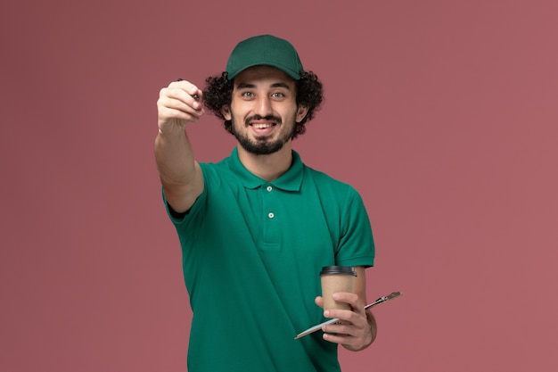 Vue avant de courrier masculin en uniforme vert et cape tenant la tasse de café de livraison avec bloc-notes et stylo sur le bureau rose service de livraison uniforme