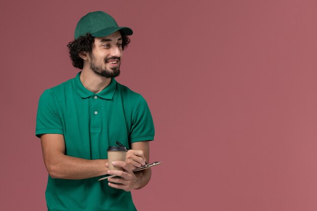 Vue avant de courrier masculin en uniforme vert et cape tenant la tasse de café de livraison avec bloc-notes et stylo sur le travail de l'entreprise de service de livraison uniforme fond rose