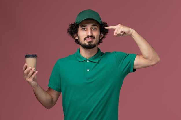 Vue avant de courrier masculin en uniforme vert et cape tenant la tasse de café de livraison sur le fond rose entreprise de services de livraison uniforme