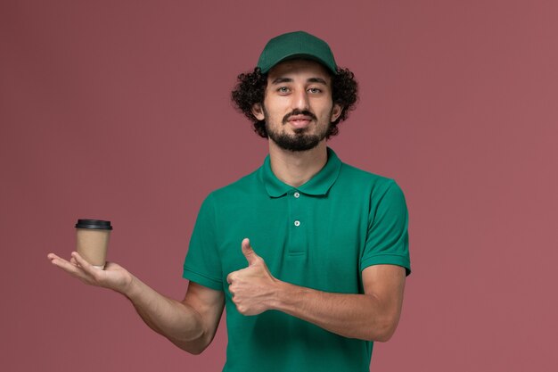 Vue avant de courrier masculin en uniforme vert et cape tenant la tasse de café de livraison sur le fond rose service de livraison uniforme travailleur de l'emploi