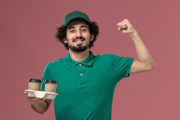 Vue avant de courrier masculin en uniforme vert et cape tenant des tasses de café fléchissant sur le fond rose service de livraison uniforme travailleur de l'emploi