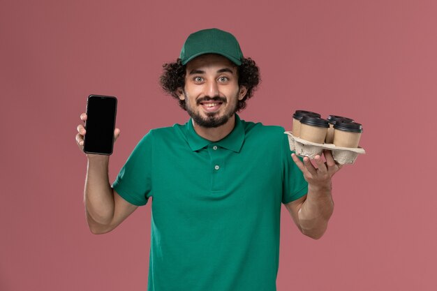 Vue avant de courrier masculin en uniforme vert et cape tenant des tasses à café avec téléphone sur le travail de livraison uniforme de service de fond rose clair