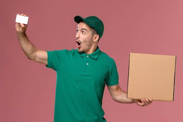 Vue avant de courrier masculin en uniforme vert tenant la boîte de nourriture avec carte blanche sur l'uniforme de travailleur de service de fond rose
