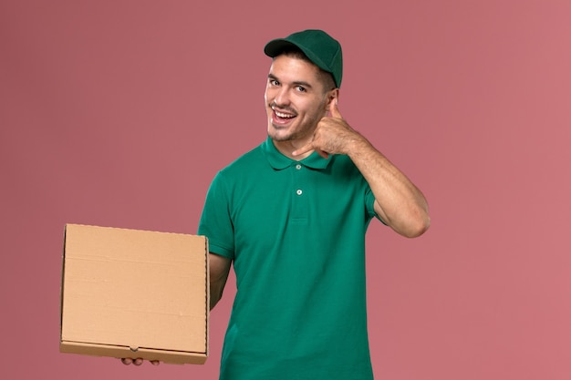 Vue avant de courrier masculin en uniforme vert tenant la boîte de nourriture et montrant l'appel téléphonique pose sur fond rose