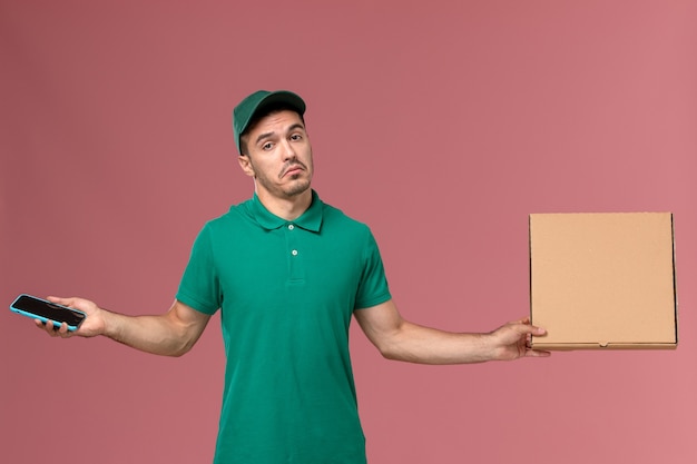 Vue avant de courrier masculin en uniforme vert tenant la boîte de nourriture et le téléphone sur fond rose