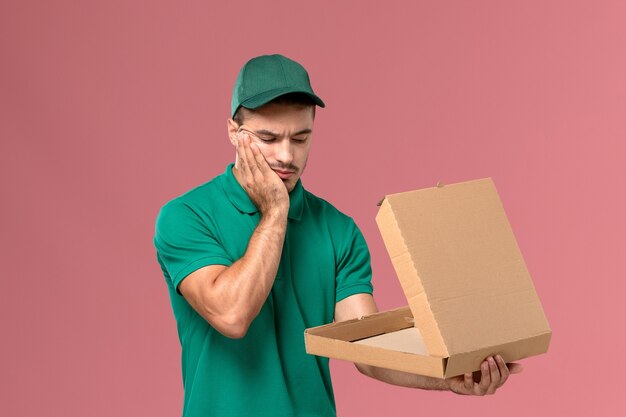 Vue avant de courrier masculin en uniforme vert tenant et ouvrant la boîte de nourriture avec une expression stressée sur fond rose clair