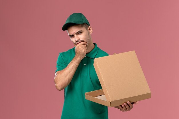 Vue avant de courrier masculin en uniforme vert tenant et ouvrant la boîte de nourriture tout en pensant sur fond rose