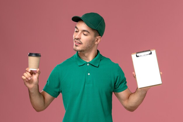 Vue avant de courrier masculin en uniforme vert tenant la tasse de café de livraison et le bloc-notes sur le bureau rose
