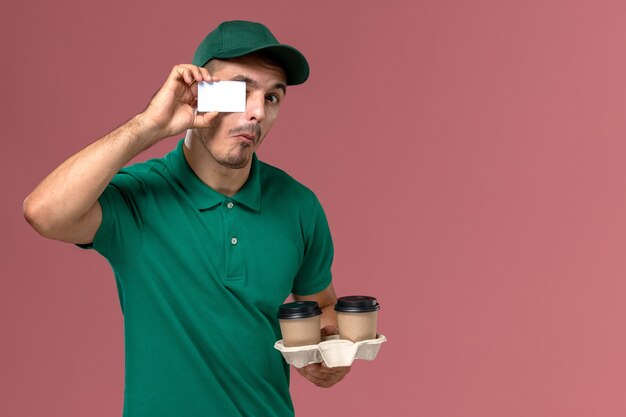 Vue avant de courrier masculin en uniforme vert tenant des tasses de café de livraison marron et carte en plastique blanc sur le bureau rose