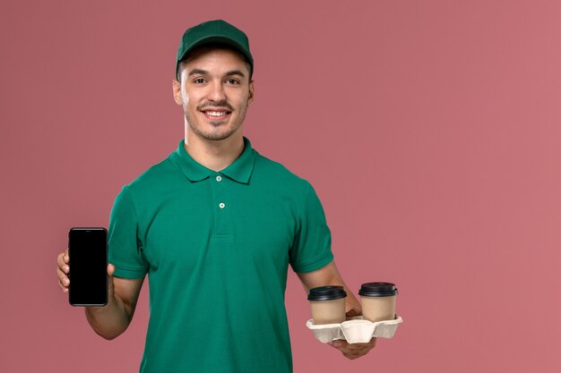 Vue avant de courrier masculin en uniforme vert tenant des tasses de café de livraison marron et téléphone sur le bureau rose