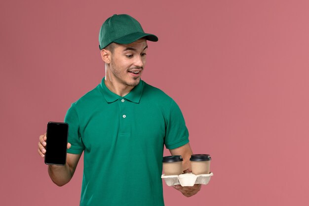 Vue avant de courrier masculin en uniforme vert tenant des tasses de café de livraison marron et téléphone sur fond rose mâle