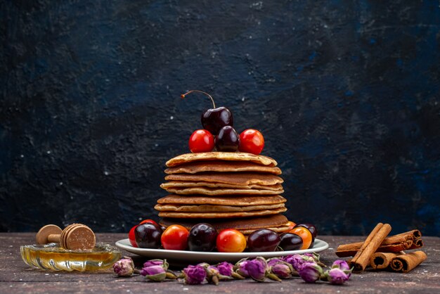 Une vue avant de délicieuses crêpes aux cerises à l'intérieur de la plaque blanche avec des fleurs et de la cannelle sur la crêpe aux fruits de bureau noir