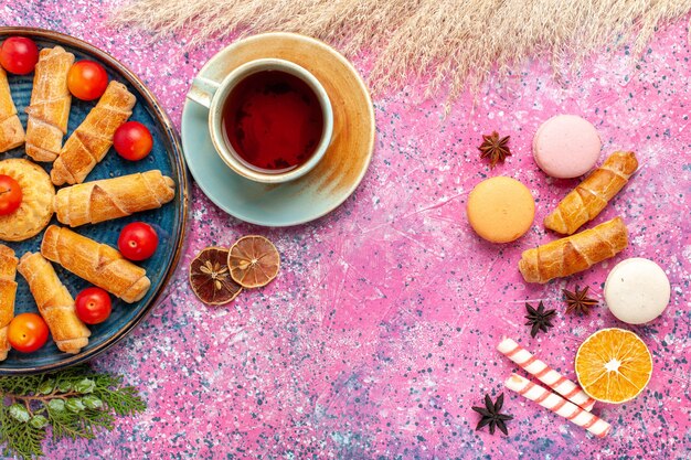 Vue avant de délicieux bagels sucrés avec des macarons français et une tasse de thé sur un bureau rose clair