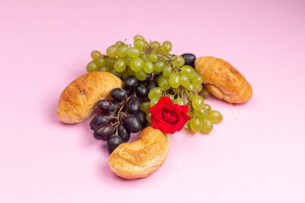 Vue avant de délicieux croissants cuits au four avec garniture de fruits avec des raisins noirs et verts frais sur le bureau rose