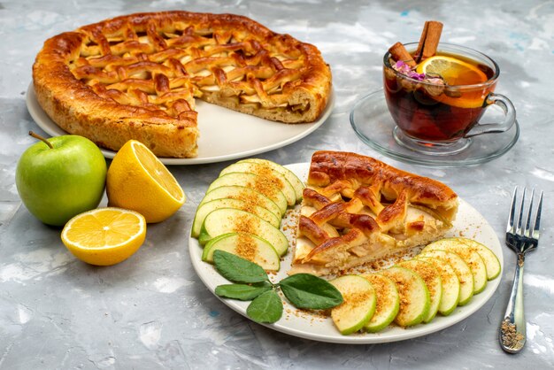Une vue avant de délicieux gâteau aux pommes à l'intérieur de la plaque avec du thé et de la pomme verte fraîche sur le sucre biscuit gâteau de bureau en bois