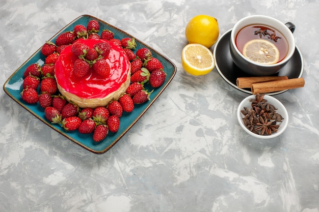 Vue avant de délicieux gâteau à la petite tarte avec une tasse de thé et des fraises fraîches sur une surface blanche