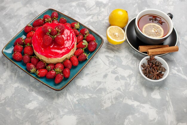 Vue avant de délicieux gâteau à la petite tarte avec une tasse de thé et des fraises fraîches sur une surface blanche