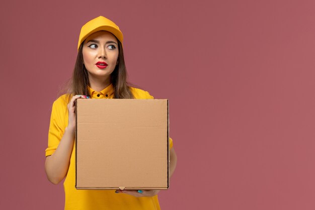 Vue avant du courrier féminin en uniforme jaune et cap tenant la boîte de nourriture sur le mur rose clair