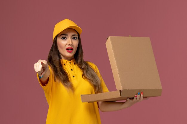 Vue avant du courrier féminin en uniforme jaune et cap tenant la boîte de nourriture ouverte sur le mur rose clair
